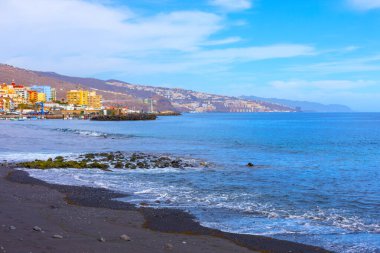 İspanya 'nın Kanarya Adaları' ndaki Tenerife adasındaki Candelaria kasabasındaki kara kum plajı. Karanlık volkanik kum, bir dizi canlı bina ve Atlantik Okyanusu 'nun arka planında yükselen dağlar.