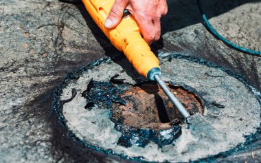 İşçi betonu kırmak için döner çekiç matkabı kullanıyor ya da asfalt kullanarak elektrik erişim noktasını kuşatıyor. İnsan beton yüzeyi kırmak için elektrikli alet kullanır. Hasarlı, katran benzeri bir alanda iş yapılıyor.