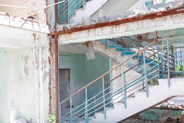 Terk edilmiş binanın içinde çürüyen beton merdiven, mavi metal parmaklıklar ve ciddi yapısal hasarlar var. Boya ve döküntüler terkedilmiş yapının her tarafına saçılmış.