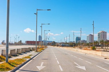 Güneşli bir günde Barcelona 'daki Platja de la Nova Mar Bella plajına giden boş bir yol. Yolun yanında şerit işaretleri ve beton bariyerler var. Arka planda modern binalar görünüyor.