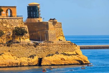 Valletta, Malta 'daki tarihi Grand Harbour' da antik taş tahkimatları ve bir dalgakıran bulunuyor. Birkaç küçük geleneksel Malta teknesi (dghajsas olarak adlandırılır) berrak mavi sularda hareket etmektedir.