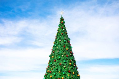 Mavi gökyüzünün altında yükselen Noel ağacı kırmızı, yeşil, gümüş ve altın süslemelerle süslenmiş. Parlak yıldız, neşeli konik şeklini taçlandırarak bayram neşesini yakalıyor.