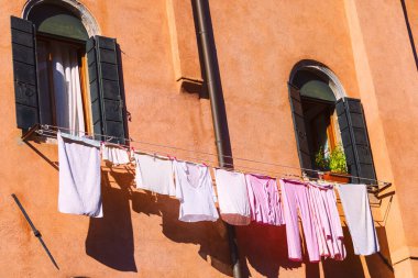 Renkli binanın dış tarafındaki kemerli pencerelerin altında asılı duran çamaşır ipinde çamaşır kurutma. Beyaz ve pembe elbiseler güneş ışığıyla aydınlanan turuncu duvara doğru seriliyor.