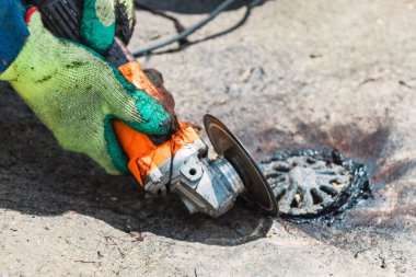 Eldiven işçisi beton öğütücüyle metal bir drenaj kapağını kesiyor. Endüstriyel bakım ve onarım işleri
