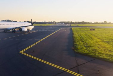 Yolcu uçağı piste iniyor, kalkışa hazırlanıyor. Sabah güneşi, asfalt ve çevreleyen yeşil çimlerin üzerine ılık bir ışık saçıyor.