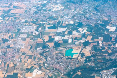 Turkuaz göl kenarındaki tarlaların ve kentsel mahallelerin havadan görünüşü. Virajlı yollar tarlaları ve kasabaları birbirine bağlıyor, toprak kullanım şekillerini ve modern manzara üzerindeki insan etkisini gösteriyor
