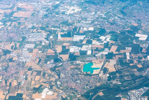 Turkuaz göl kenarındaki tarlaların ve kentsel mahallelerin havadan görünüşü. Virajlı yollar tarlaları ve kasabaları birbirine bağlıyor, toprak kullanım şekillerini ve modern manzara üzerindeki insan etkisini gösteriyor
