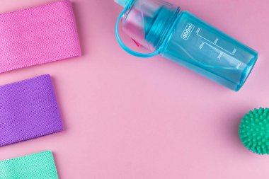 Pembe arka planda renkli fitness dişetleri. Elastik genişleticiler ve farklı renkte kasetler. Renkli lastik ve tekstil bantları, bir şişe su ve masaj miyofasyal topu. Evde spor aletleri.