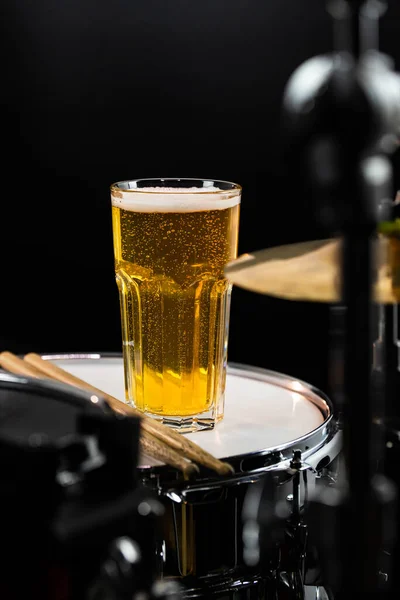 Glass of light beer with salty snacks on professional drum set closeup. Drumsticks, drums and cymbals, at live music rock concert, in the club stage, bar, or in recording studio. Black background.