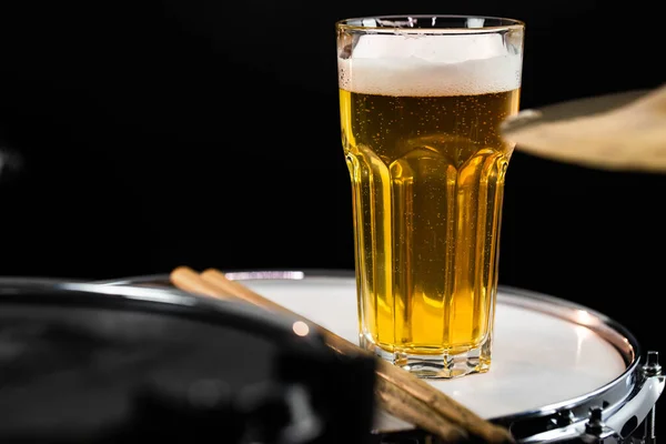 Glass of light beer with salty snacks on professional drum set closeup. Drumsticks, drums and cymbals, at live music rock concert, in the club stage, bar, or in recording studio. Black background.