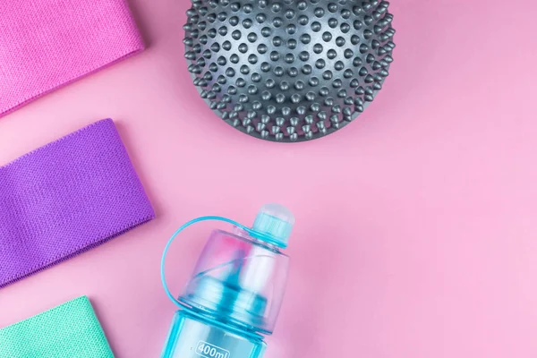 Colorful fitness gums on pink background. Elastic expanders and tapes of different color. Colored rubber and textile bands, bottle of water and massage myofascial ball. Home fitness equipment.