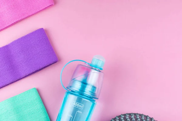 Colorful fitness gums on pink background. Elastic expanders and tapes of different color. Colored rubber and textile bands, bottle of water and massage myofascial ball. Home fitness equipment.