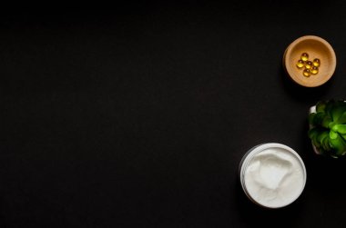 Kozmetik kremli beyaz kap, ahşap bir çanakta vitaminli kapsüller, siyah arka planda kapalı bir çiçek. Mesaj için yer. Düz Yatay
