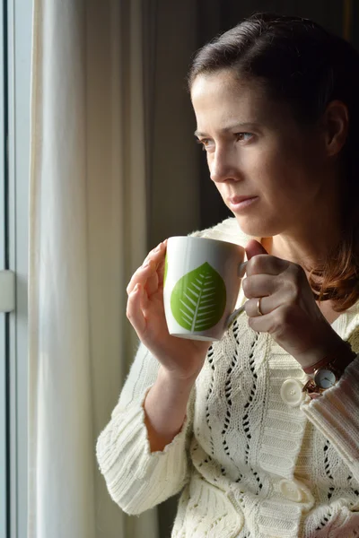 Woman holds a mug looks out of a window — ストック写真