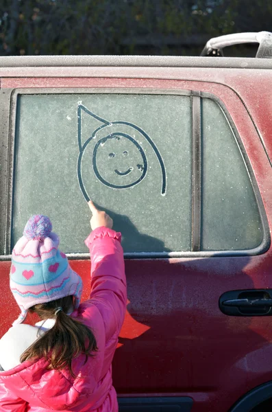 小さな女の子が霜で覆われた車の窓を幸せそうな顔を描く — ストック写真