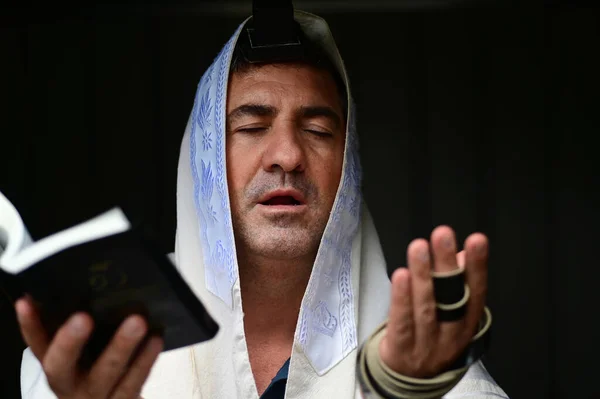 Adult Jewish man holding Siddur book (Jewish prayer book) , wearing tallit and tefillin (Jewish prayer clothing), paying the morning pray.Isolated on black background. Real people. Copy space