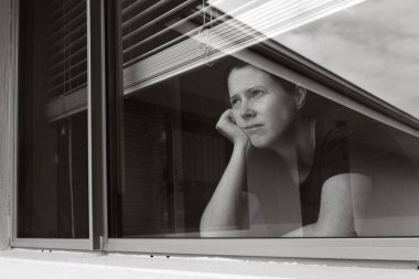 Sad adult woman (age 30-40) looking out home window glass. Real people. Copy space clipart