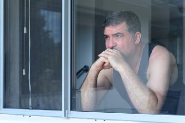 Orta yaşlı üzgün bir adam (40-50 yaş) ev penceresinden bakıyor. Gerçek insanlar. Boşluğu kopyala