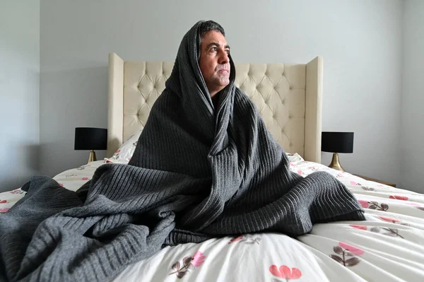 Afraid adult man (age 40-50) covered in blanket sitting on bed in bedroom. Mental illness concept.Real people. Copy space