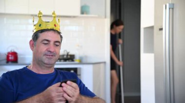 Tembel yetişkin bir adam (40-50 yaş) kral gibi davranıyor, karısı çalışırken ve mutfağı temizlerken cep telefonuna mesaj atıyor. Tembellik konsepti. Gerçek insanlar. Boşluğu kopyala