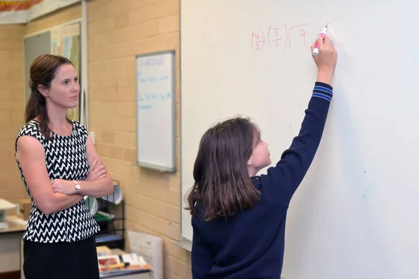 高中女生 30岁至40岁的女教师 看着一名女生 12岁至13岁 在学校教室的白板上解答一个数学问题 — 图库照片