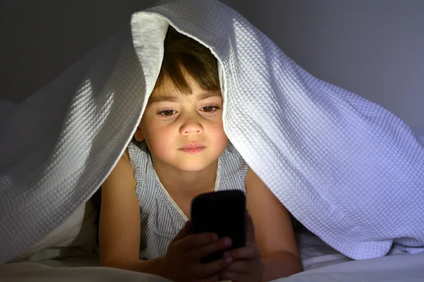 Маленький ребенок играет на смартфоне в постели под одеялом в почти — стоковое фото