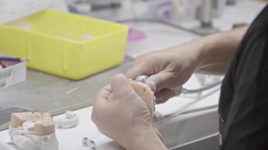 Laboratuvarda hassas aletlerle takma diş modelini şekillendiren bir diş teknisyeninin yakın çekimi. Profesyonel dişçi üretim süreci işçiliği ve tıbbi imalatı gösteriyor.