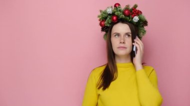 Geleneksel Noel çelengi giymiş genç bir kadın cep telefonuyla konuşuyor hoş olmayan konuşmalar yapıyor, pembe stüdyo arka planında kopyalanmış bir şekilde fotokopi çekiyor. Mutlu yıllar, mutlu yıllar.