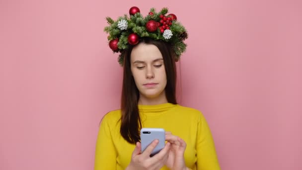 かなり若い女性は あなたの広告コンテンツのためのコピースペース パステルピンクのスタジオの壁の上に隔離された快適な会話を行う携帯電話で話して クリスマスリースを身に着けています 明けましておめでとうございます — ストック動画