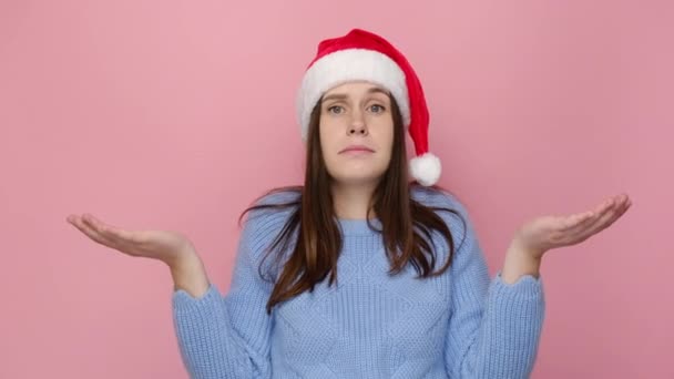 困惑した表情で赤いクリスマスの帽子の若い女性が混乱した表情で手を広げ 困惑し ピンクのスタジオの背景の壁に隔離されたスタンド お正月お祝いメリーホリデー — ストック動画