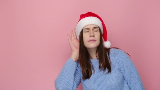 クリスマスの赤い帽子のかわいい若い女性は ピンクの背景スタジオに隔離された居心地の良い青いセーターを着て あなたが熱心に耳を傾けることを耳にしようとします ハッピーニューイヤーお祝いメリーホリデーコンセプト — ストック動画