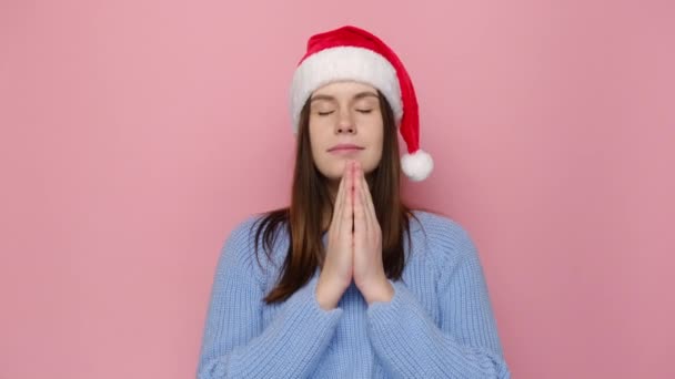 クリスマスの赤い帽子で満足の良い外観の女性は 祈りのジェスチャーでヤシの木を保持し 肯定的な表現 穏やかな笑顔をして 願いを行い ピンクのスタジオの壁に隔離された青いセーターを着ています クリスマスが来る — ストック動画