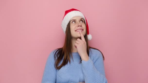 クリスマスの帽子の夢のような魅力的な若い女の子は ピンクのスタジオの背景に隔離された居心地の良いセーターを着て 思慮深い表現を考えています お正月お祝いメリーホリデー — ストック動画