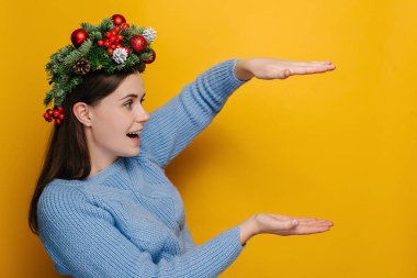 Shocked young woman in Christmas wreath showing something large of big size looking at hands, amazed girl with open mouth surprised with huge discounts gesturing, isolated on yellow studio background clipart
