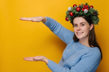 El yapımı Noel çelengi içinde gülümseyen genç bir kadın dikey fotokopi aletiyle boy gösteriyor, sarı stüdyo arka planında izole edilmiş. Mutlu yıllar 2021 bayram partisi konsepti