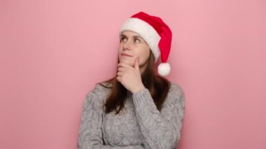 Pembe stüdyo arka planında izole edilmiş düşünceli, kafası karışık genç bir kadın yeni fikirle işaret parmağını kaldırdı kışlık rahat süveter ve Noel şapkası giydi. Yeni yıl kutlamaları, mutlu tatiller.