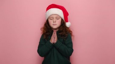 Sakin şirin küçük kız avuç içlerini birbirine bastırır Noel Baba 'nın kırmızı şapkası ve kış süveteri giyer pembe stüdyo arka planında modeller yapar. Yalvarma ve mutlu yıllar konsepti