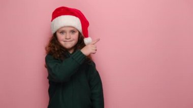 Şirin küçük kız, reklam içeriği için ön parmağı açık, Noel şapkası ve süveteri giymiş, pembe arka planda izole edilmiş. Yeni yılı kutluyoruz. Mutlu tatiller.
