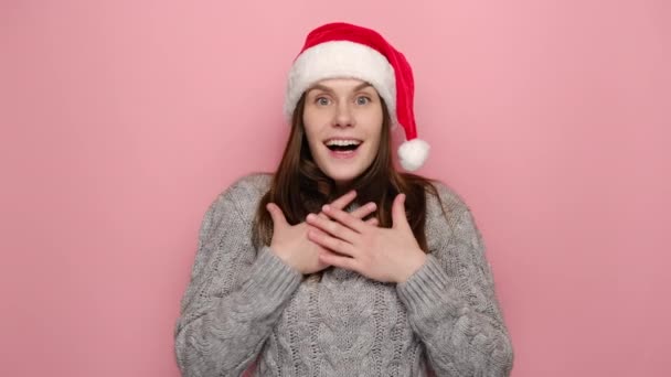 ショックを受けた表情でサンタクリスマスの帽子の幸せな笑顔の若い女性は頬に手のひらを保ち 冬の居心地の良いセーターを着て 新年を待ち ピンクのスタジオの背景上のモデル クリスマスが来る — ストック動画