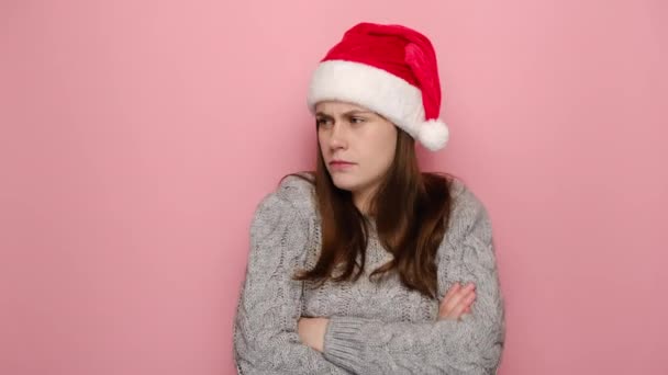 胸の上に折り畳まれた交差手で立ってカメラを見てクリスマスの帽子で悲しいいらいらした若い女性の肖像画は ピンクのスタジオの壁に隔離された冬のセーターを着ています お正月の陽気な休日のコンセプト — ストック動画