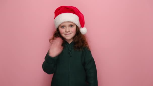 クリスマスの赤い帽子で幸せな笑顔の女の子の子供 こんにちはジェスチャーで波のヤシの木 友人を迎え セーターを着て ピンクのスタジオの背景上のモデル ハッピーニューイヤーお祝いメリーホリデーコンセプト — ストック動画