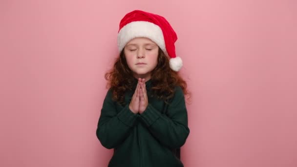 冷静可爱的小女孩让手掌紧握在一起祈祷 穿着圣诞红帽子和冬季毛衣 模特们穿着粉色工作室的背景 祈求与新年快乐的概念 — 图库视频影像