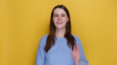 Cheerful young caucasian woman 20s pointing index fingers on camera doing phone gesture like says call me back blinking, dressed in sweater, isolated on yellow background. People lifestyle concept