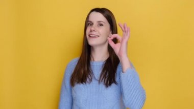 Smiling funny young caucasian woman 20s looking camera showing Ok gesture blinking, dressed in cozy blue sweater, posing isolated on yellow studio background. Emotion people lifestyle concept.