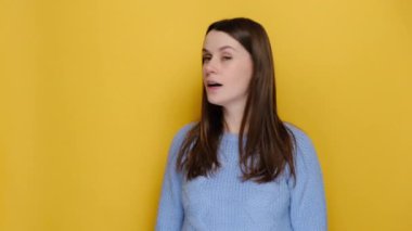 Shocked amazed young caucasian woman 20s pointing index finger aside say wow, locking at camera, wears casual blue sweater, isolated on yellow background in studio. Emotion people lifestyle concept.