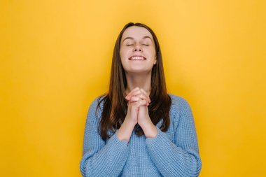 Umut dolu genç bir kadın yumruklarını sıkıyor, dilek diliyor, hayallerin gerçekleşmesi için dua ediyor, umutlu bir kız, iyi şansa inanıyor, süveter giyiyor, sarı stüdyo arka planında izole edilmiş.