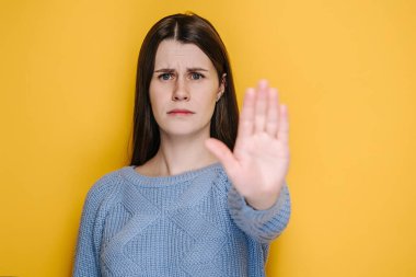 Kendine güveni tam genç bir bayan dur hareketi yapıyor, esmer bir kız kameraya bakıyor düşüncesizce telkin etmeyi reddediyor ya da teklif ediyor sarı stüdyo arka planında izole edilmiş mavi kazak giyiyor.