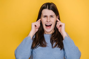Kafkasyalı çılgın genç kadın portresi kulaklarını kapatıyor parmakları sürekli çığlık atıyor, mavi kazak giyiyor, sarı stüdyo arka planında izole edilmiş poz veriyor. Duyguların insanları yaşam tarzı konsepti