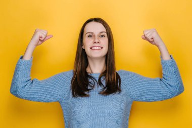 Çekici genç bir kadın kaslarını gösteriyor ve kendine güveniyor gibi görünüyor, kadın hakları için savaşmak için güç hissediyor, sarı stüdyo arka planında izole edilmiş mavi süveter giyiyor. Ben güçlü ve bağımsızım.!