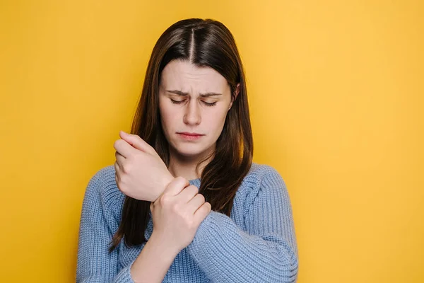 厌倦了不健康的年轻女性按摩疼痛的手 患有腕管综合症 穿着蓝色毛衣 模特在黄色工作室的背景 扭伤腕部 肌肉僵硬 — 图库照片